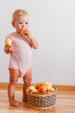 Sevimli gülümseyen bir yaşındaki kız yalın ayak ayakta duruyor ve beyaz arka planda olgun armutları yiyor.