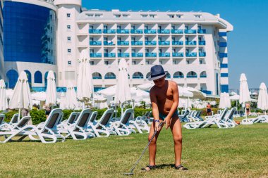 Bronzlaşmış 9 yaşındaki şapkalı çocuk yazın otel bahçesinde mini golf oynuyor..