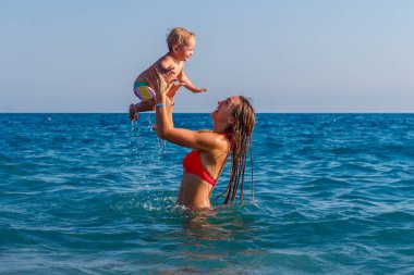 Genç, uzun saçlı, gülümseyen bir kadın ve yazın denizde bir yaşında bir kız bebek..