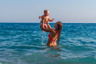 Genç, uzun saçlı, gülümseyen bir kadın ve yazın denizde bir yaşında bir kız bebek..