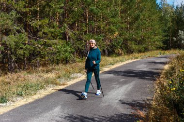 60 yaşında spor giyim ve spor ayakkabı giyen mutlu bir kadın İskandinav yürüyüşü için direklerle pistte yürüyor.