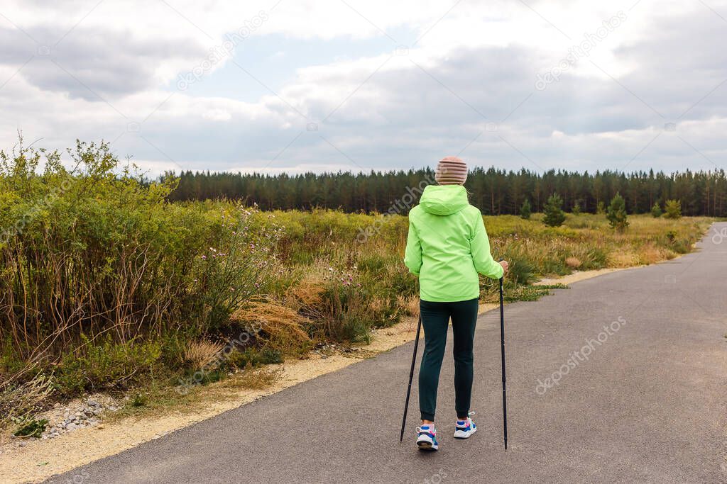 a woman in a light green windbreaker with Poles for Nordic walking stands with her back outdoors