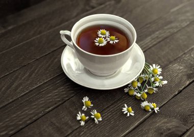 Beyaz bir porselen çay fincanı ve çay tabağı papatya çayıyla doludur. Her yerde papatya çiçekleri var..