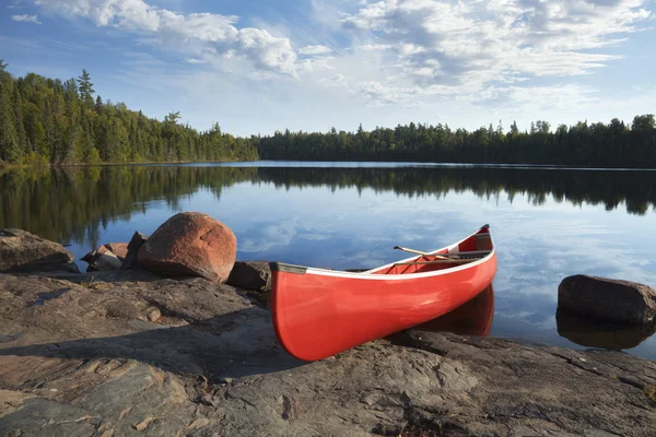 Red canoe on rocky shore of calm lake with pine trees — Stock Photo, Image