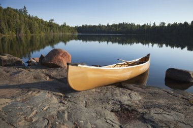 Yellow canoe on rocky shore of calm lake with pine trees clipart