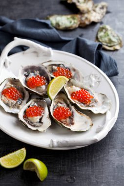 Kırmızı havyar ve limonla süslenmiş taze istiridye. Beyaz bir tabakta servis edilmiş. Gri beton arkaplan