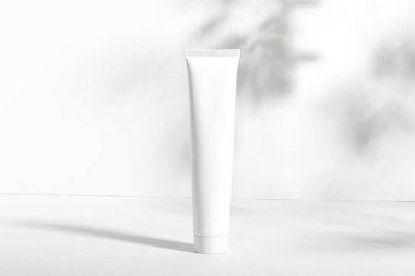 Beyaz arka planda, yaprak gölgeli beyaz bir kozmetik tüp. Besleyici ve nemlendirici yüz kremi, el kremi ya da losyon. Cilt bakım ürünüyle kadınların kozmetik tüpü. Organik kozmetik ürünleri, kopyalama alanı