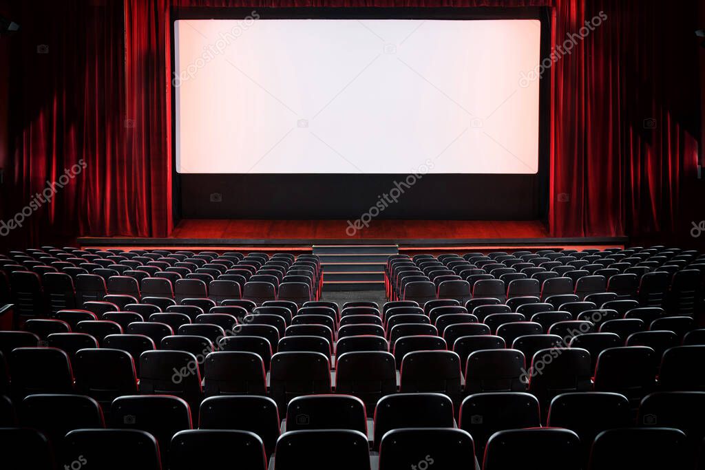 Auditorium of an empty movie theatre and stage with opened red velvet curtains viewed from the rear of the rows seats with lights on the screen