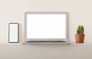 Akıllı telefon ve dizüstü bilgisayar beyaz boş ekran ve kaktüsle aydınlık duvara yaslanmış vaziyette.