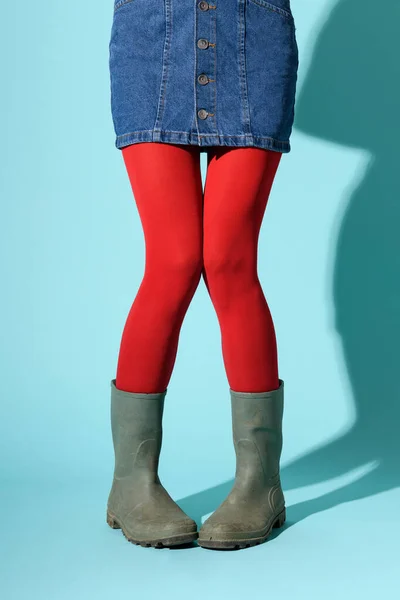 Funny legs of a young woman in denim skirt, red collant and green rubber boots posing with knock knees and pigeon toes over a blue background with copyspace