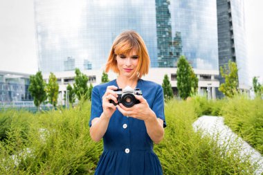 Kamerasını şehirdeki kadın turist, modern cam gökdelenin arka planına karşı çekimini yaparken sessiz bir gülümsemeyle kamerayı işaret ediyor.