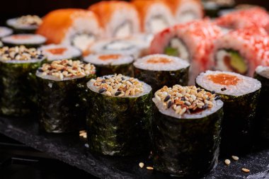 Dilimlenmiş suşi rulolarının üzerine sushi tohumları serpiştirilmiş diğer suşi rulolarının arka planındaki siyah tahta üzerinde. Japon Doğu mutfağı..
