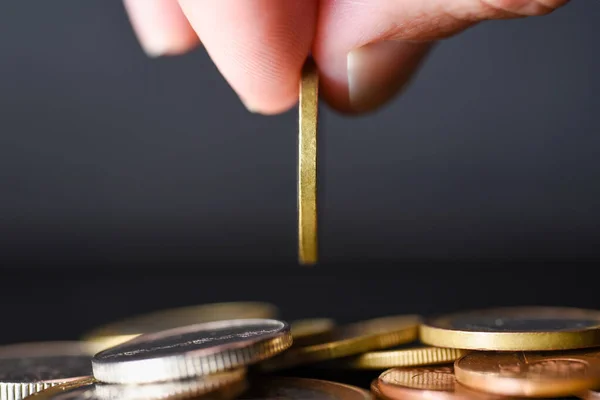 Hand drops a coin into a stack of coins on a black background.