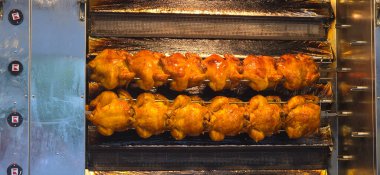 Kızarmış tavuklar, ticari bir mutfaktaki bir rostoda yemek pişiriyor. Altın-kahverengi bir ten ve yoğun bir mutfak atmosferi sergiliyorlar..