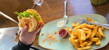 Elinde taze bir hamburger, yanında marul ve sos var. Yanında da kırsal bir ahşap masada patates kızartması ve sos var..