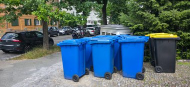 Berlin, Almanya - 3 Temmuz 2025: Cadde boyunca park halindeki arabaları ve yemyeşil çimenlikleri olan renkli çöp kutuları yerleştirildi.