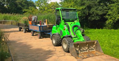 Berlin, Germany - September 08, 2025: Green compact tractor is hauling a trailer filled with garden tools along a scenic pathway.