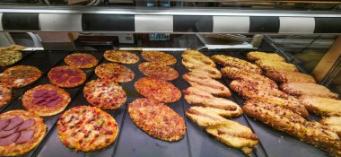 Çeşitli pizzalar ve ekmek çubukları yeni pişmiş ve fırında sergileniyor. Müşteriler için çeşitli malzemeler ve kabuklar sergileniyor..