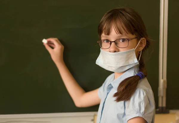 Adorable student in mask is writing on the chalkboard in classroom. Close-up portrait of little girl holding a piece of chalk during Covid-19 pandemic