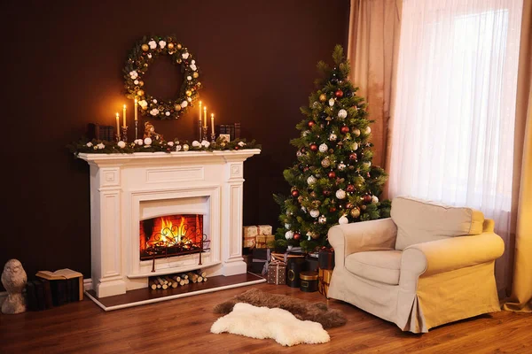 Dark living room interior with comfortable armchair next to the fireplace decorated with a wreath with lights and candeles. Christmas tree, gifts, and pillows. Xmas Home Interior Decoration