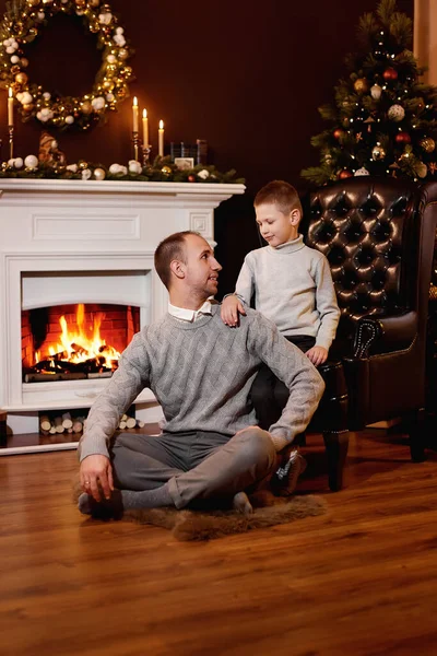 Father and son sitting on floor near the fireplace and the Christmas tree, looking at each other. Beautiful leather armchair, New Year\'s wreath on the wall. Candles burn on the fireplace and books.