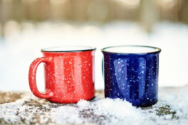 Bright red and blue mugs with hot drinks on the snow. Background with text space for your promotion or website about holiday weekend during the cold season. Winter holiday activity concept, Christmas clipart