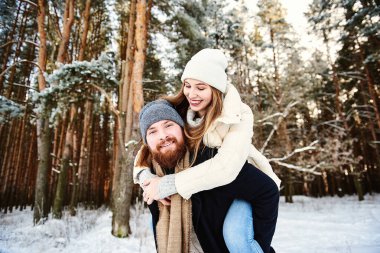 Gülümseyen mutlu bir çift. Erkek, karlı ormanda kış tatilinde kadını sırtında taşıyor. Genç bir çift kış ormanında yürüyor. Noel zamanı. Kış tatili romantik ilişki konsepti