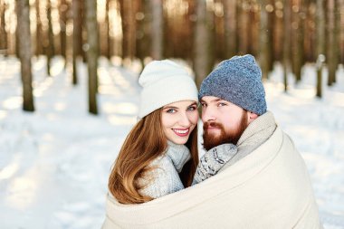 Kameraya bakan gülümseyen bir çiftin yakın plan portresi, karlı kış gününde açık havada yürümenin keyfini çıkarıyor. Arkadaki ormanda güzel bir kış günbatımı, Sevgililer Günü. Birlikte Noel tatili