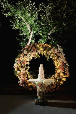 Akşam düğün pastası. Bir çiçek kavisinin arka planında pasta. Krem ve dekorasyonun beyaz ve pembe renkleri. Farklı boyutlarda dört kat. Gece düğünü.