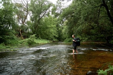Ekose gömlekli ve yeşil şortlu büyük bir sırt çantalı bir adam nehrin uzak kıyısına bakarak nehri geçer. Yürüyüş konsepti. Ekipman, yürüyüş için teçhizat. Metin için boşluk