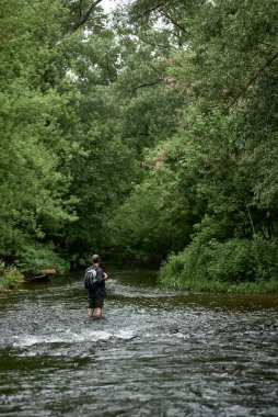 Ekose gömlekli ve sırt çantalı bir erkek yürüyüşçünün nehri geçip doğanın tadını çıkarışını. Ekipmanlar, yürüyüş ekipmanları, seyahat tutkusu yaşam tarzı, vahşi doğa konseptinde tatiller. Dikey fotoğraf