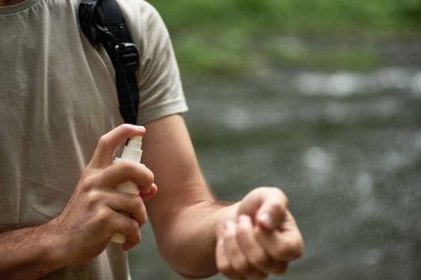 Nehir kenarındaki ormanda yürüyüş yaparken sivrisinek ve kene püskürtücü kullanan erkek turist, cilt koruması uyguluyor, seçici bir odak noktası var.