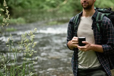 Nehrin kıyısında duran büyük bir sırt çantalı yürüyüşçü ve elinde sıcak içecekle siyah bir bardak tutuyor. Kamp gezileri, gezgin yaşam tarzı..