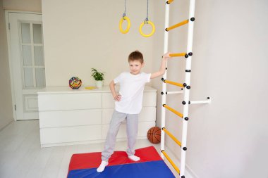 Çocuk kameranın önünde poz veriyor, çerçeveye bakıyor. Evin spor köşesi. Spor, sağlıklı yaşam tarzı, evde boş vakit geçirme. Mutlu küçük çocuk evde spor yapıyor.