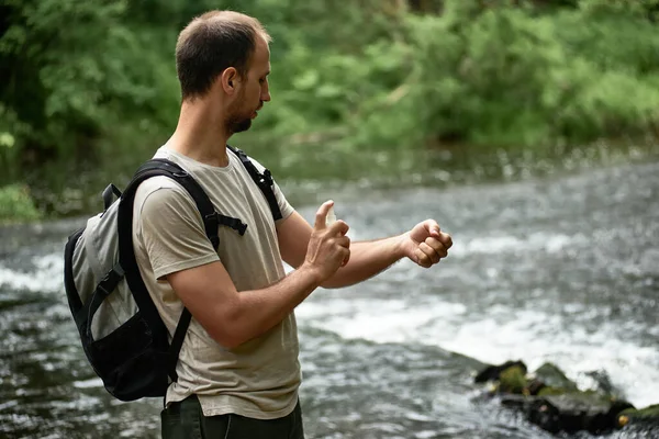 Side view of a hiker in a shirt with a backpack near the river spraying insect repellent on skin. Equipment, gear for hiking,camping essentials. Wanderlust lifestyle, vacations in the wilderness.