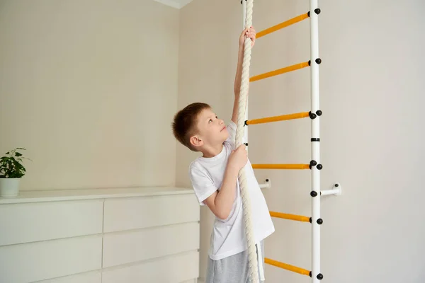 Evde tırmanma aktivitesi. Beyaz tişört ve gri eşofman giyen yakışıklı okul çocuğu evde beden eğitimi sırasında ayaklarını bar duvarına dayayıp yukarı bakıyor, ipe tırmanıyor.