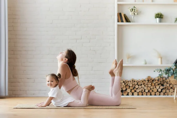 可爱的小女孩看着相机笑着 在妈妈做了半个弓形瑜伽后重复她的身体柔韧运动 母女俩都在家里健身 — 图库照片