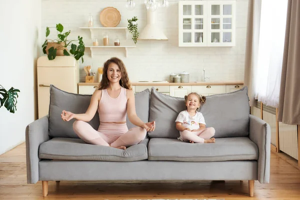 笑着活跃的年轻妈妈和小女儿坐在沙发上练习莲花式瑜伽 快乐有趣的妈妈和女孩用泥巴手冥想 过着健康的生活 — 图库照片