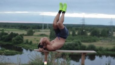 Parlak spor ayakkabıları içinde çıplak bir gövdesi olan genç bir vücut geliştirici doğada antrenman yapar ve bir bankta uzanırken karın kaslarını sallar. Yan görüş. Yakın plan.