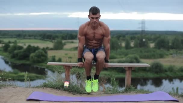 Mavi şortlu genç bir atlet nehir kenarındaki bir bankta amuda kalkıyor ve bacaklarını bükerek baskıyı sallıyor. Ön manzara. Yakın plan. — Stok video