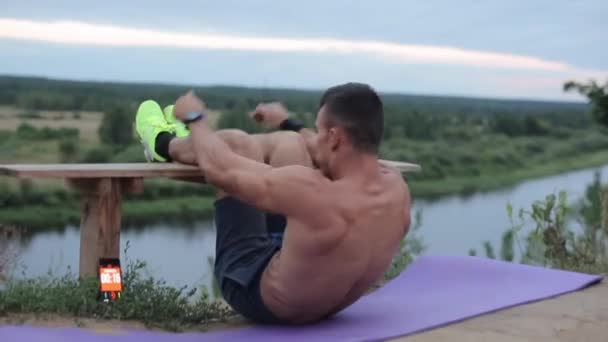 Un joven culturista trabaja junto al río y bombea sus abdominales encendiendo el temporizador de intervalos en su teléfono. Primer plano. La cámara sigue al sujeto — Vídeos de Stock