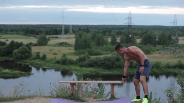 A young guy in blue shorts is resting after a set of strength exercises and restoring his breathing by putting his phone on a bench by the river. Medium plan — Stock Video