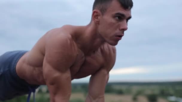 Un jeune bodybuilder avec un torse nu s'entraînant dans la nature et fait des pompes sur un banc au bord de la rivière. Gros plan. Tir au niveau des yeux. La caméra se déplace de haut en bas — Video