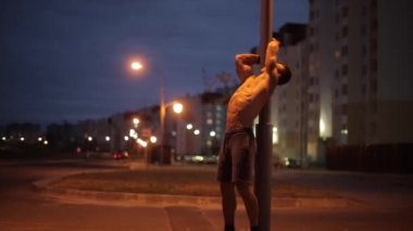 Mavi şortlu genç bir adam şehirde bir yaz akşamı antrenman yapıyor ve sokak lambasına insan bayrağı dikiyor. Yan görüş. Orta boy. Bulanık arkaplan