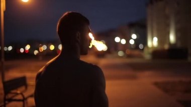 Çıplak gövdeli genç bir sporcu şehrin arka planına karşı duruyor ve etrafına bakıyor. Arkadan bak. Yakın plan. Kamera soldan sağa hareket ediyor.