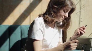 Misk, Belarus - 25 Ağustos 2020: Beyaz tişörtlü enerjik genç bir kız kafedeki bir masada oturuyor ve elinde kozmetik bir kalem tutarken aynaya bakıyor. Yan görüş. Yakın plan.