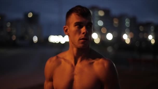 En ung muskulös man utan t-shirt träning på torget och vilar mellan övningar medan du står mot bakgrund av kvällens stadsljus. Framifrån. Närbild. suddig bakgrund — Stockvideo