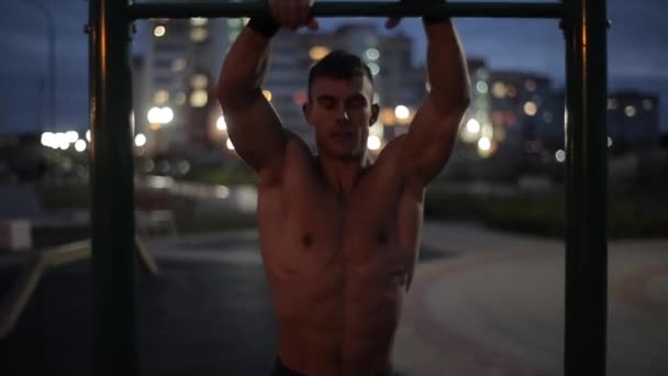 Ein junger Bodybuilder ohne Hemd trainiert an einem Sommerabend auf einem städtischen Sportplatz und zieht an einer waagerechten Stange hoch. Frontansicht. Nahaufnahme. Unklarer Hintergrund — Stockvideo