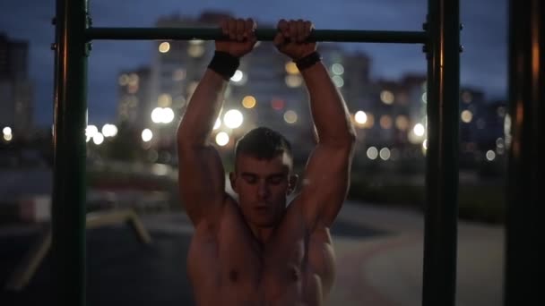 Mladý silný sportovec s nahým trupem dělá na vodorovné tyči zatáčky na pozadí rozmazaných světel večerního města. Přední pohled. Detailní záběr — Stock video