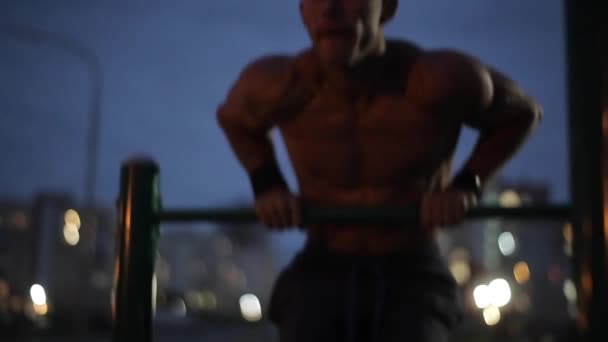 A young strong athlete with a naked torso does pull-ups on the horizontal bar against the background of blurry lights of the evening city. Front view. Close-up — Stock Video